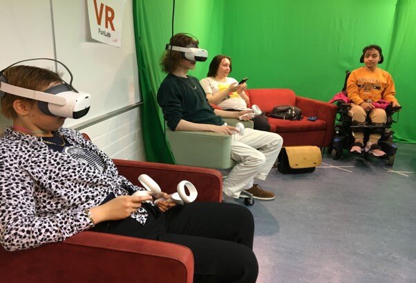 Etualalla olevat mies ja nainen istuvat sohvatuoleilla VR-lasit silmillä. Taaempana mies ja nainen seuraavat tilannetta. Huoneen takaseinällä on iso vihreä green screen kangas.